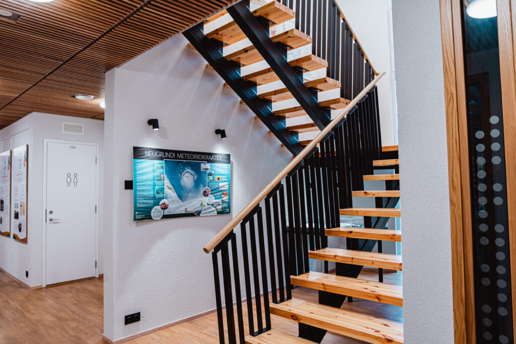 Moodne interjöör avatud puittrepi ja mustade metallpiiretega. Seinal infotahvel Neugrundi meteoriidikraatri kohta ning kõrval koridor valge ukse ja puidust põrandaga.