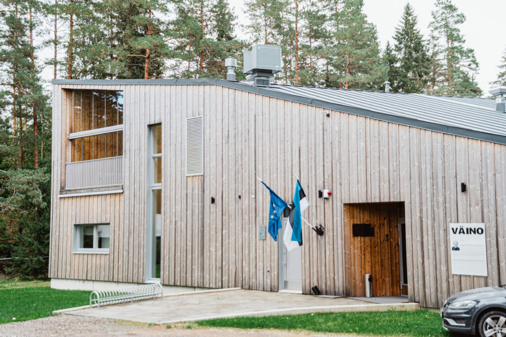 Moodne puidust fassaadiga hoone nimega „Väino“, mille ees lehvivad Eesti ja Euroopa Liidu lipud. Esiplaanil jalgrattahoidla ja parkiv auto, taustal männimets.