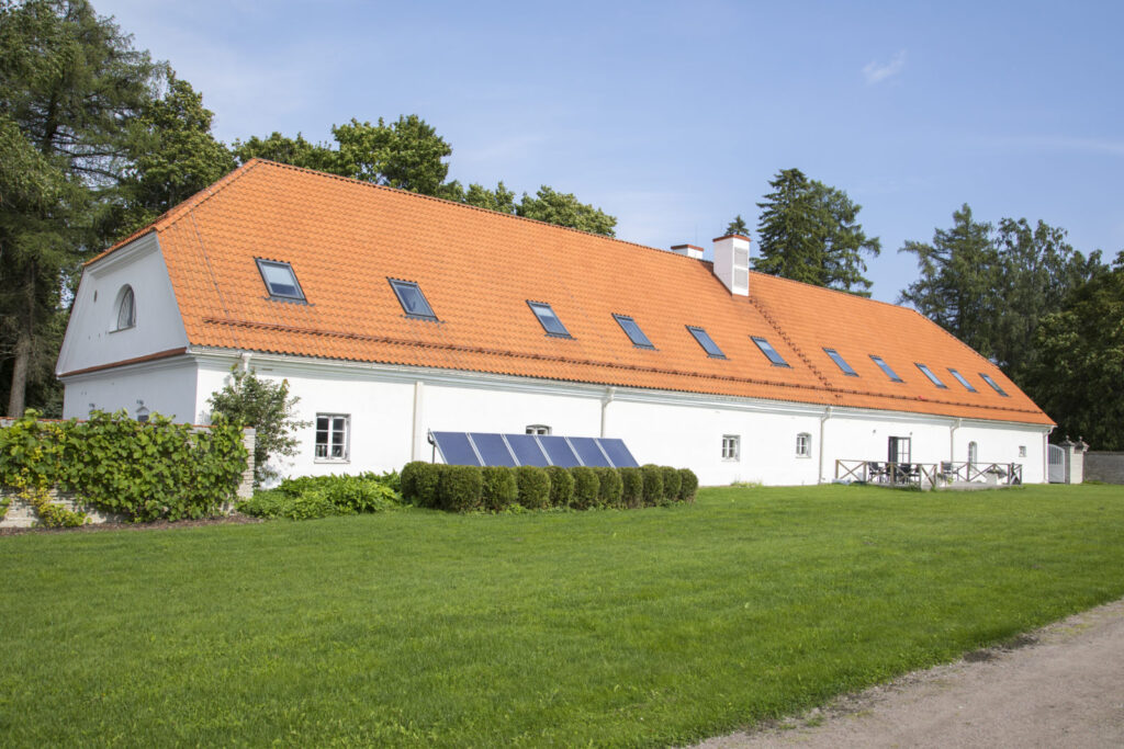 Valge pikliku hoone vaade punase kivikatuse ja katuseakendega, esiplaanil hoolitsetud muru ja päikesepaneelid, taustal rohelised puud ja sinine taevas.