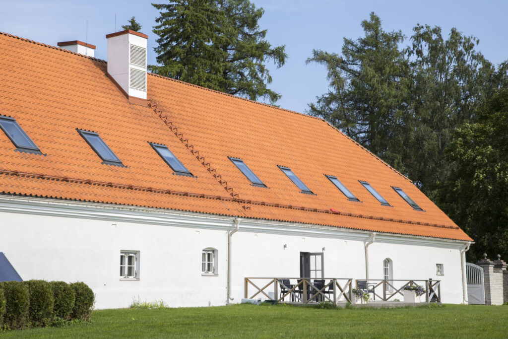 Valge kivimaja oranži viilkatusega, millel on rida katuseaknaid; ees roheline muru ja terrass toolide ning laudadega, taustal kõrged puud.