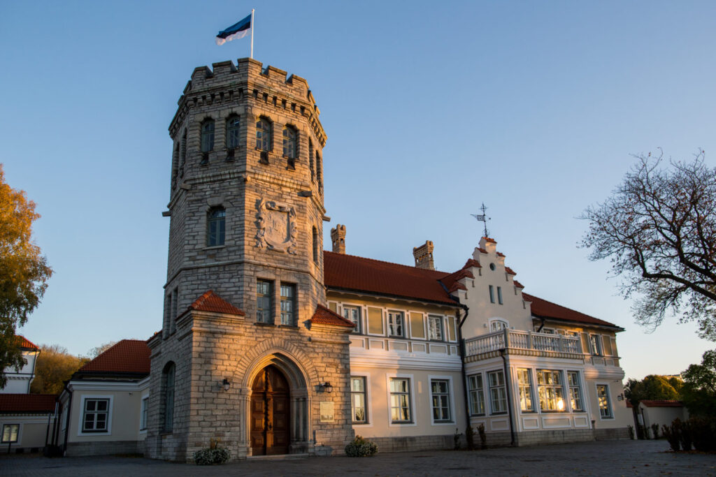 Eesti Ajaloomuuseumi Maarjamäe loss õhtupäikeses – hele paekivist torn Eesti lipuga, kõrvale jääb renoveeritud peahoone punase katuse ja dekoratiivse fassaadiga.