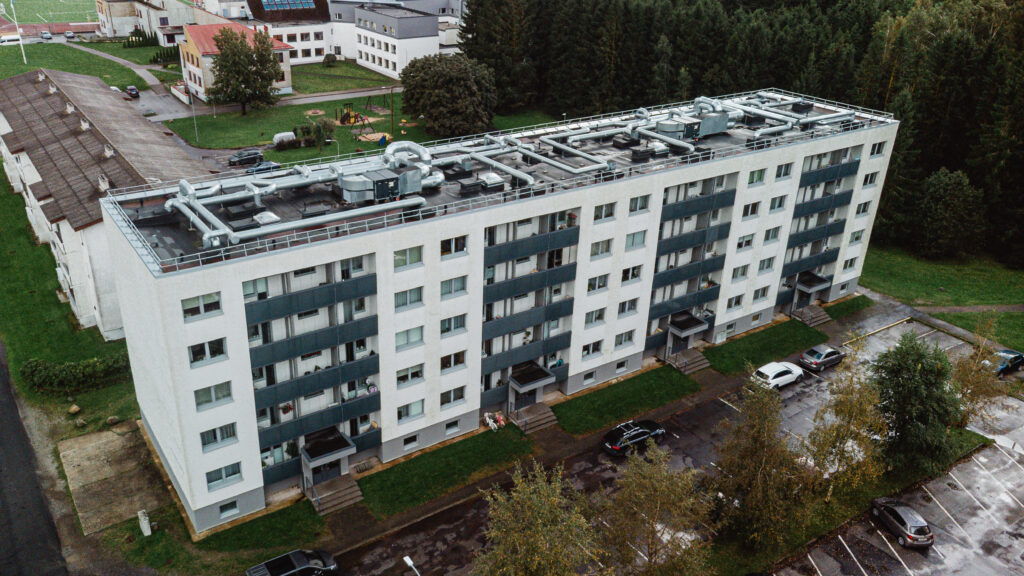 Droonifoto heleda fassaadiga kuuekorruselisest kortermajast hallide rõdude ja ventilatsiooniseadmetega katusel, ees parkimisala ja taustal haljasala ning teised elamud.
