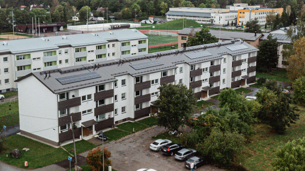 Droonivaade renoveeritud korterelamule Sügise 2, mille katusel on päikesepaneelid; taustal Aruküla staadion ja koolihooned.