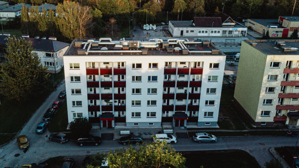 Linnavaade droonilt, esiplaanil viiekorruseline korterelamu punaste rõdude ja heleda fassaadiga, mille ees on pargitud autod ja tagaplaanil näha hooneid ja puid.