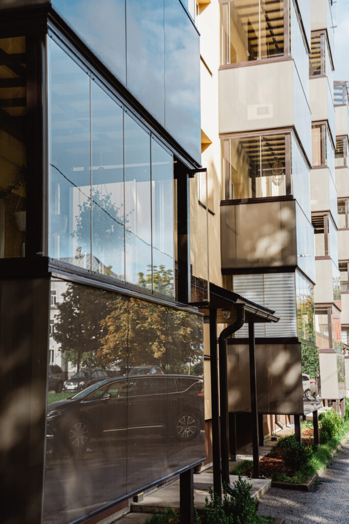Klaasitud rõdudega renoveeritud korterelamu detailvaade, rõdu peegeldustes on nähtavad puud ja tänaval parkivad autod.