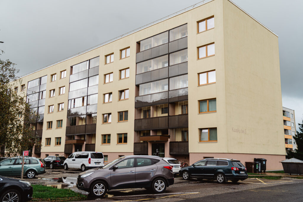 Kuuekorruseline helebeeži fassaadiga kortermaja pruunide klaasitud rõdudega ja parklaga maja ees.