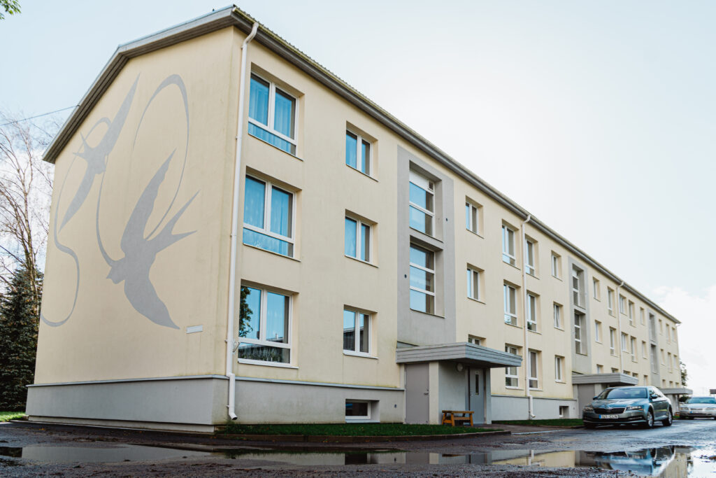 Helekollane kolmekorruseline korterelamu Rakvere mnt 6, mille otsaseinal on kujutatud lendavaid pääsukesi; hoone ees seisab auto ja peegeldus on näha vihmalombis.